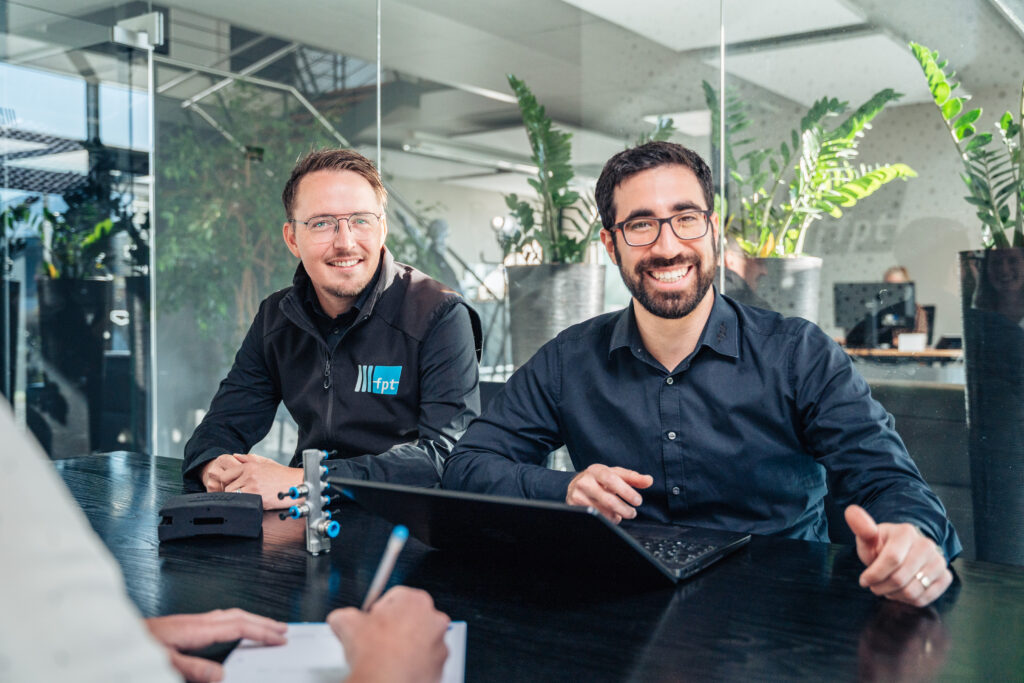 Besprechungssituation in einem lichtdurchfluteten Besprechungszimmer zwischen zwei Mitarbeitern, einer davon am Laptop