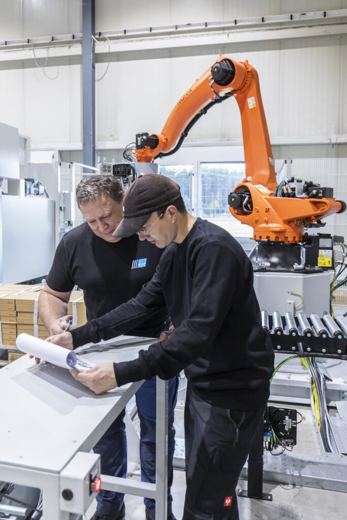 Service employee at the plant talking to construction plans and drawings, an orange 6-axis robot in the background
