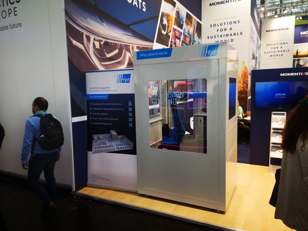 Robotic cell next to a roll-up banner at an exhibition stand, with a person walking by