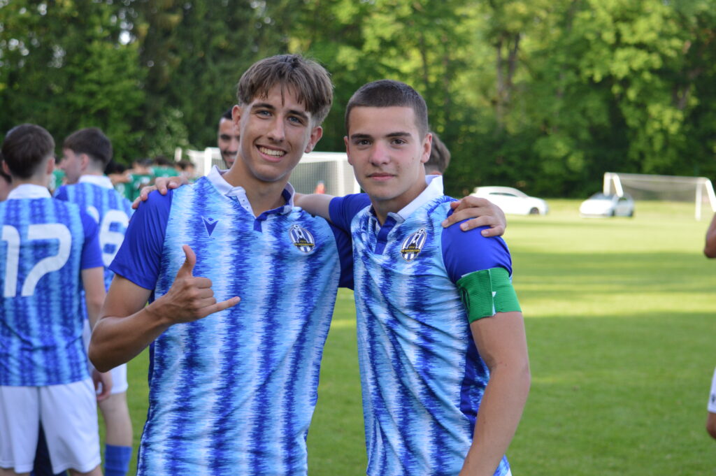 Zwei Jungs stehen Arm in Arm vor der Kamera und lächeln. Beide in Fußballtrikots.