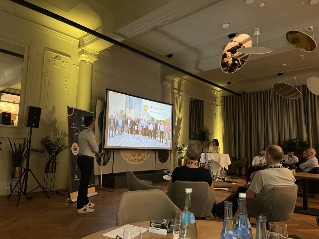 Ein in gelb beleuchteter Saal, in dem eine Präsentation stattfindet, der Menschen beiwohnen