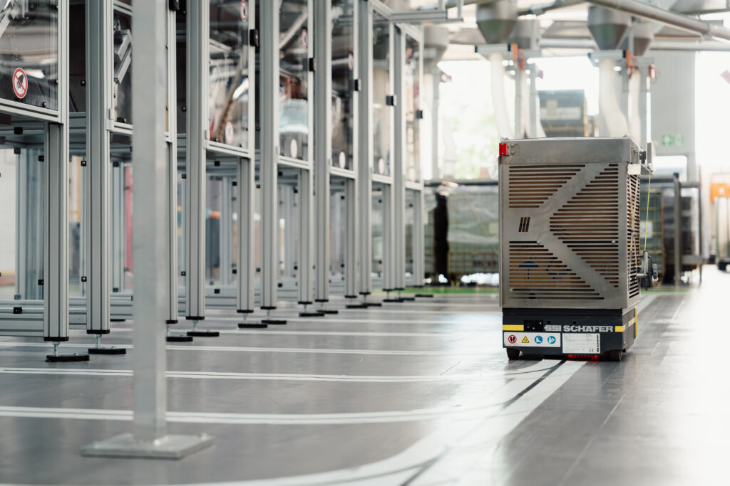 Fahrerloses Transportsystem mit Aufschrift SSI Schäfer und einem metallischen Aufbau fährt auf Lininen durch die Produktion