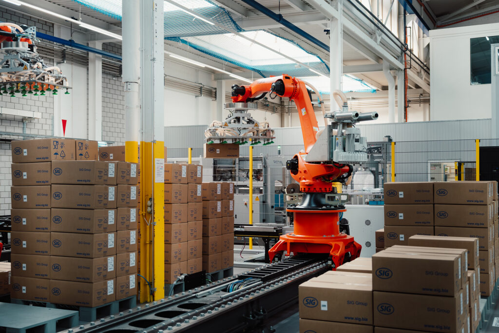 Ein orangener Roboter neben mehreren Paletten von gleichartigen aufgestapelten Kartons in einer Produktionshalle