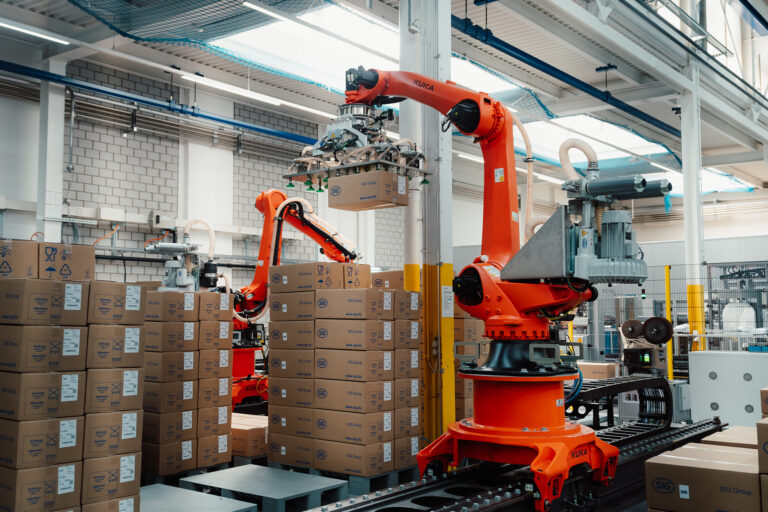 Ein orangener Roboter mit Vakuum-Sauggreifer palettiert ein Karton auf bereits bestückte Paletten