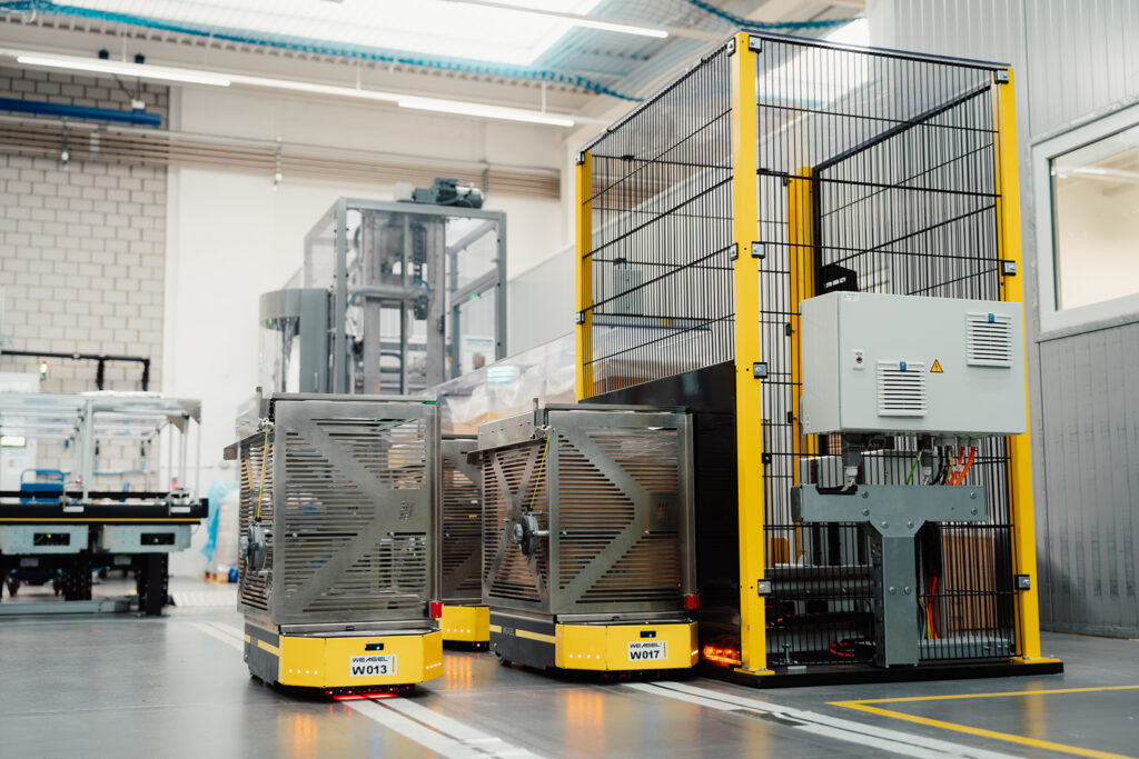 In einer Produktionsumgebung navigieren fahrerlose Transportsysteme in Gelb mit metallischem Aufsatz entlang von weißen Fahrspuren, daneben ein Schutzzaun mit Öffnung